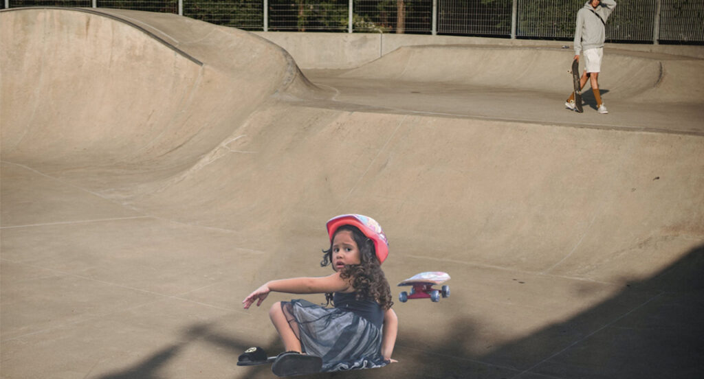 A young girl with a pained expression is on the ground with a skateboard nearby as an older skateboarder is in the distance with a hand on his head