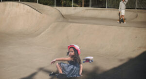A young girl with a pained expression is on the ground with a skateboard nearby as an older skateboarder is in the distance with a hand on his head